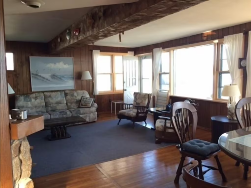 Cozy living room with wooden walls, large windows, a sofa, armchairs, and a dining table, featuring a warm, inviting atmosphere.