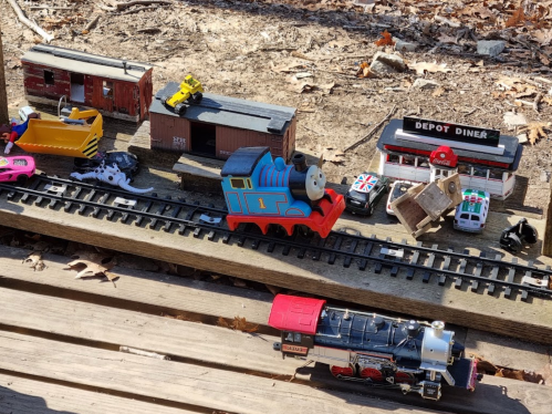 A colorful toy train on tracks, surrounded by various toy vehicles and a diner model, set on a wooden surface.
