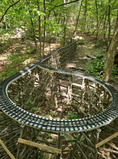 A winding model train track made of wood, set in a lush green forest with trees and foliage surrounding it.