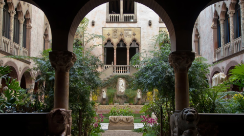 A serene courtyard with lush greenery, stone sculptures, and an archway framing a historic building in the background.