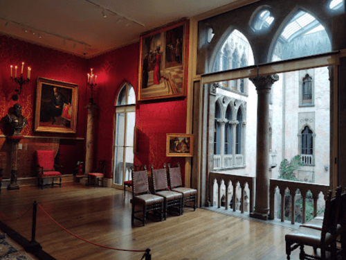 Interior of a room with red walls, paintings, wooden chairs, and a view of a balcony with gothic arches.