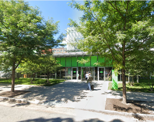 A modern building with green accents, surrounded by trees, featuring large glass doors and a clear blue sky.