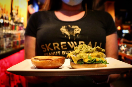 A person holds a plate with a burger topped with green toppings and a toasted bun, wearing a Skrewball Peanut Butter shirt.