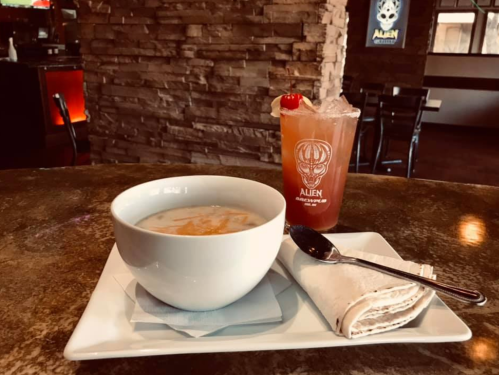 A bowl of soup with a spoon and a cocktail with a cherry, set on a stone table in a restaurant.