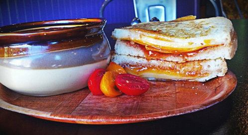 A grilled cheese sandwich with melted cheese, served with a small bowl of soup and colorful cherry tomatoes on a wooden plate.