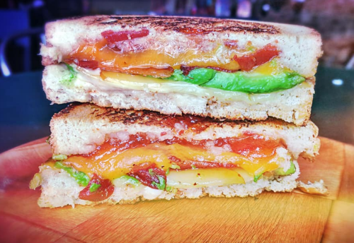 A close-up of a grilled cheese sandwich with layers of cheese, avocado, and bacon, served on a wooden plate.