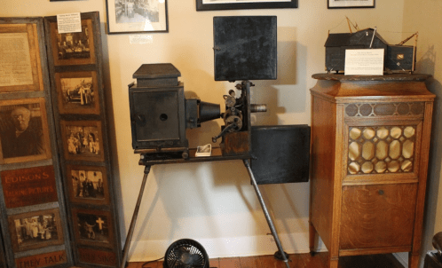 An antique film projector displayed in a museum, surrounded by historical photographs and a vintage cabinet.