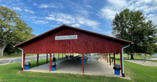 A red pavilion at Edison Park with picnic tables, surrounded by green grass and a playground in the background.