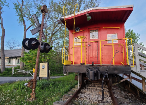A red caboose on tracks beside a vintage railroad crossing sign, surrounded by greenery and trees.