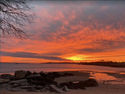 Vibrant sunset over a calm beach, with orange and pink hues reflecting on the water and rocky shoreline.