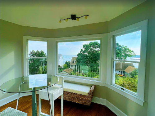 Bright room with a glass table and white chairs, featuring large windows overlooking a scenic view of houses and water.