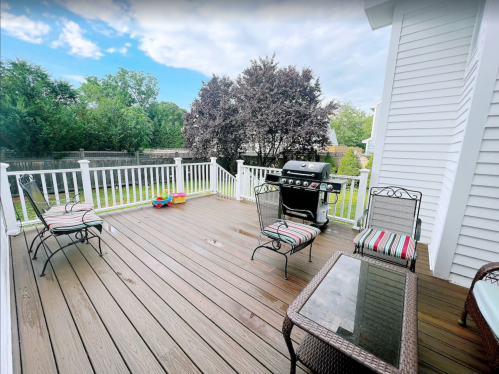 A wooden deck with two chairs, a glass table, and a grill, surrounded by greenery and a cloudy sky.