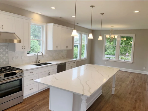 Modern kitchen with white cabinets, marble island, stainless steel appliances, and large windows overlooking greenery.
