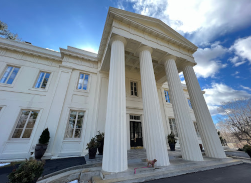A grand white mansion with tall columns and a large entrance, set against a blue sky with scattered clouds.