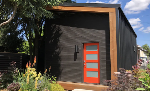 Modern black house with a vibrant orange door, surrounded by lush greenery and colorful flowers under a blue sky.