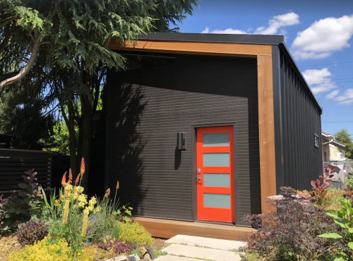 Modern black house with a wooden accent and a bright orange door, surrounded by colorful plants and blue sky.