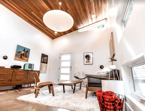 Modern living room with wooden ceiling, stylish furniture, and artwork, featuring a cozy rug and bright natural light.