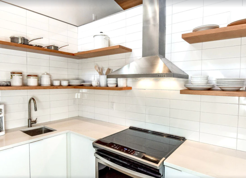 Modern kitchen with white tile walls, wooden shelves, stainless steel appliances, and a sleek countertop.