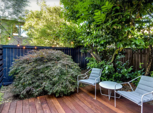 A serene garden with lush greenery, a wooden deck, two chairs, and a small table surrounded by plants.