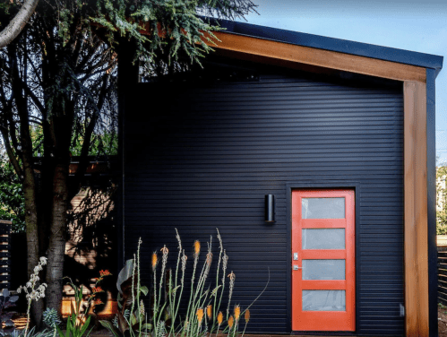 Modern black house with a wooden accent, featuring a bright orange door and surrounded by greenery.