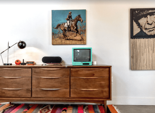 A stylish room featuring a wooden dresser, vintage TV, artwork of a horse rider, and a modern lamp on a colorful rug.