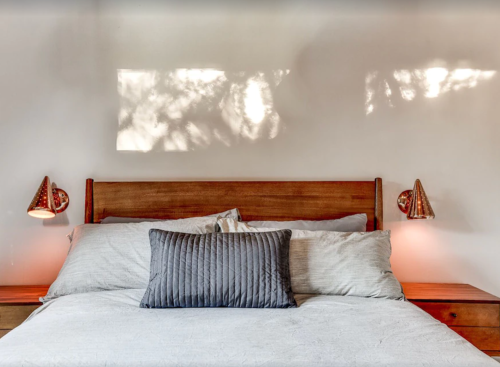 A cozy bedroom with a wooden headboard, gray bedding, and warm wall sconces casting soft shadows.