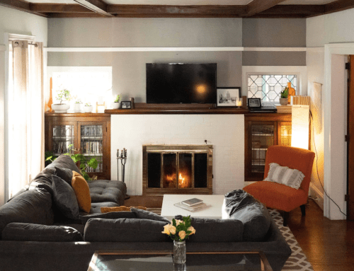 Cozy living room with a gray sectional sofa, orange chair, fireplace, and a TV on the wall, featuring plants and warm lighting.