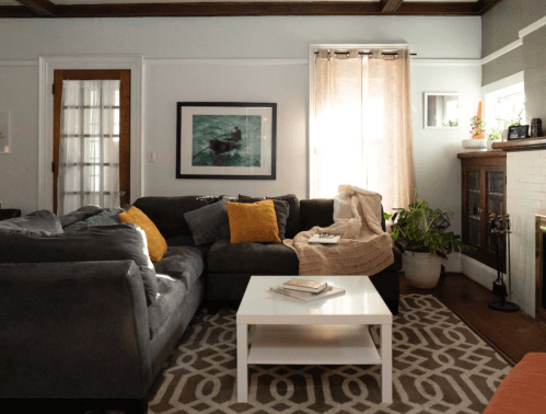 Cozy living room with a gray sectional sofa, white coffee table, and natural light from a window. Plants and decor add warmth.