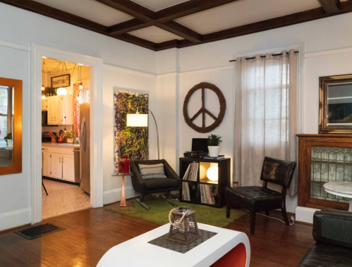 Cozy living room with wooden accents, a peace sign decor, and a view into a kitchen. Modern furniture and warm lighting.