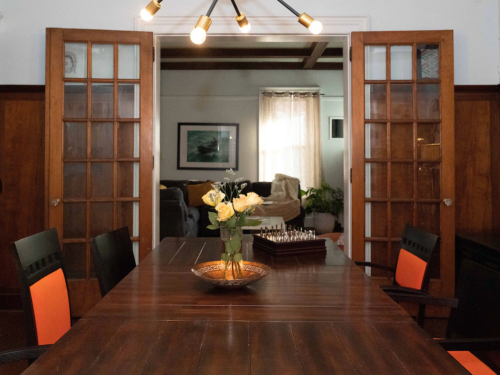 A dining table with a vase of yellow flowers, surrounded by chairs, leading to a cozy living room.