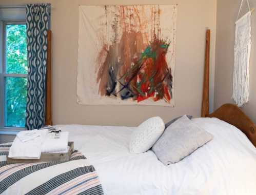 Cozy bedroom featuring a bed with pillows, a colorful wall hanging, and a tray with towels on a striped blanket.