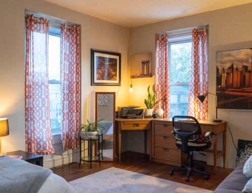 Cozy room with a bed, desk, plants, and large windows dressed in patterned curtains. Warm lighting creates a welcoming atmosphere.