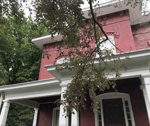 A red brick house with white trim, featuring a porch and surrounded by green trees.