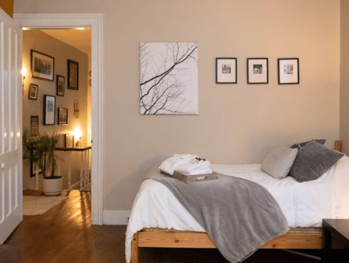 A cozy bedroom with a bed, gray blanket, and framed artwork on the wall, leading to a softly lit hallway.