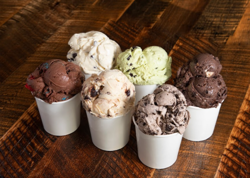 Seven cups of various ice cream flavors arranged on a wooden table, including chocolate, mint, and cookies and cream.
