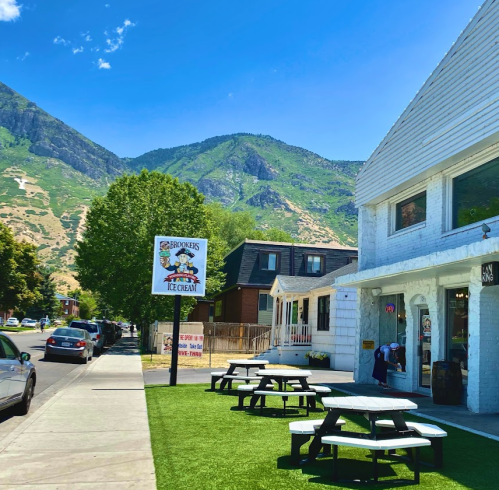 A sunny street scene featuring an ice cream shop, picnic tables, and mountains in the background.