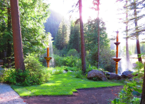 A serene forest scene with three water fountains surrounded by lush greenery and tall trees, illuminated by sunlight.