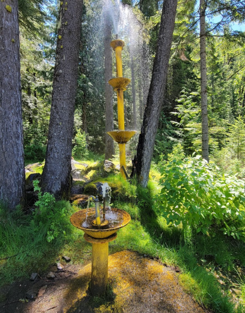 A yellow water fountain surrounded by trees and greenery, with water spraying from the top into a basin below.