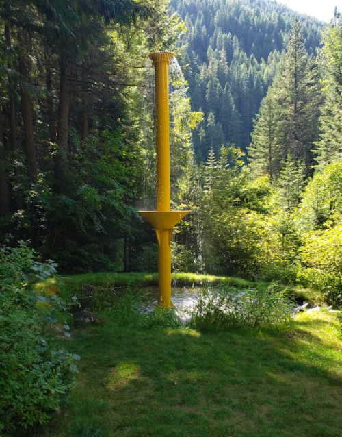 A tall yellow water feature in a lush green forest, with water cascading down into a small pond surrounded by trees.