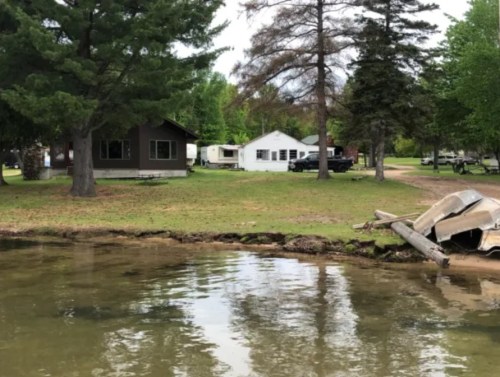 A serene lakeside view featuring two houses, a trailer, and a partially submerged boat near the shore.