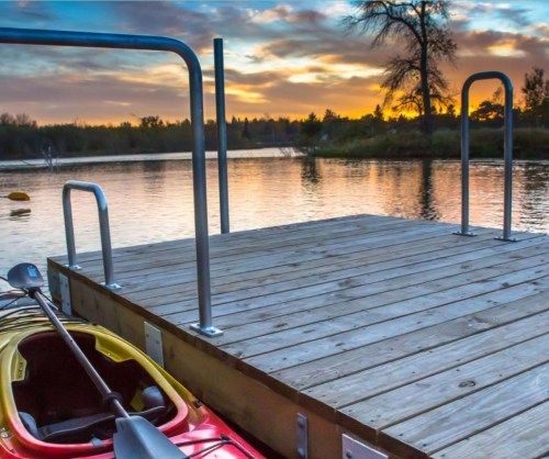 A wooden dock extends over a calm lake at sunset, with a kayak resting nearby and trees silhouetted in the background.