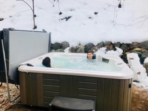 Two people relax in a hot tub surrounded by snow-covered rocks and trees.