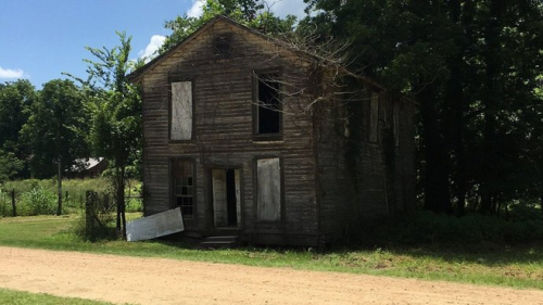 Abandoned wooden house with boarded windows, surrounded by greenery and a dirt road.