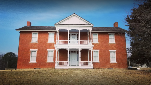 A large red brick house with a white porch and balcony, set against a clear blue sky.