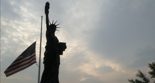 Silhouette of the Statue of Liberty against a cloudy sky, with an American flag at half-mast in the foreground.