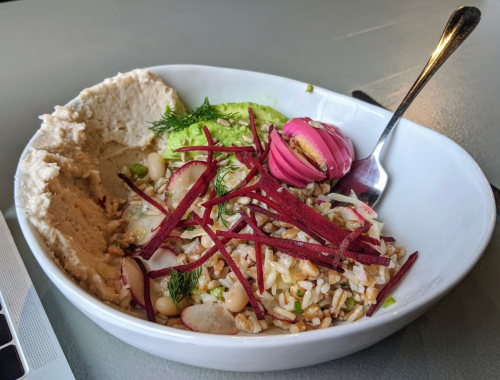A bowl of grain salad with radishes, beets, and hummus, garnished with herbs and a pink dessert on the side.