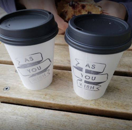 Two coffee cups with black lids, featuring the phrase "AS YOU WISH" printed on them, placed on a wooden table.