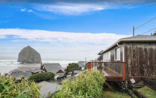 A coastal view featuring a house, ocean waves, and a large rock formation in the distance under a blue sky.