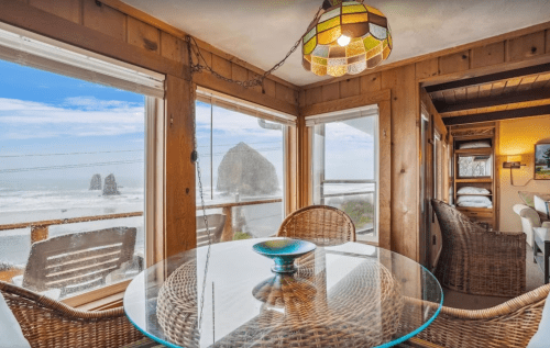 Cozy interior with a glass table, wicker chairs, and ocean views featuring large rock formations in the distance.