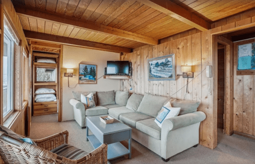 Cozy wooden cabin interior featuring a green sofa, TV, and bunk beds, with natural light from large windows.
